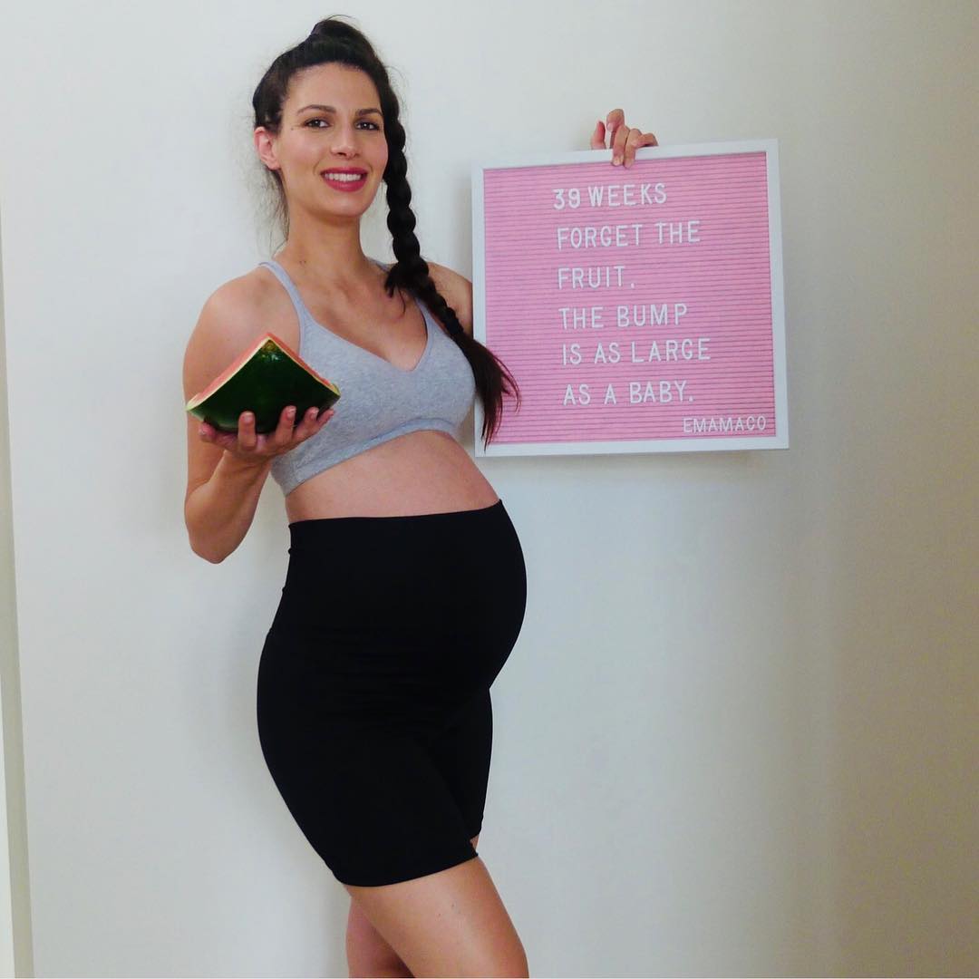 Pregnant woman in black maternity bike shorts outfit standing in a sunny park, holding her baby bump and showcasing third-trimester bump style.