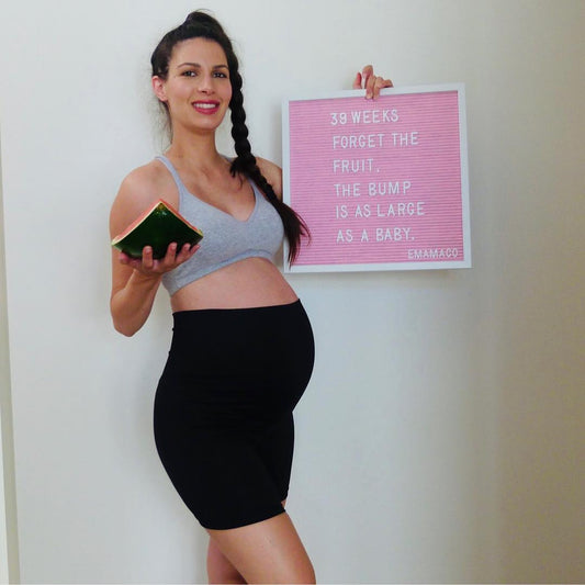 Pregnant woman in black maternity bike shorts outfit standing in a sunny park, holding her baby bump and showcasing third-trimester bump style.