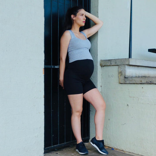 Pregnant woman in grey tank top and black body shapewear shorts leaning against an outdoor wall near a black metal gate, wearing sneakers, showing second trimester maternity shapewear shorts outfit